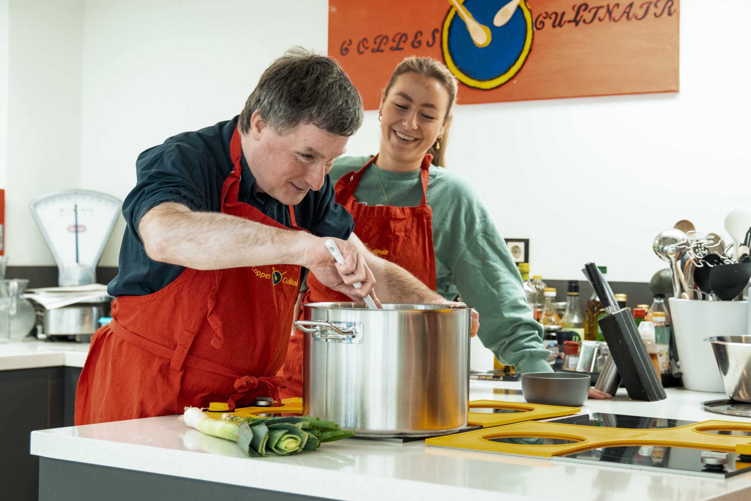 Twee personen staan in een moderne keuken en bereiden samen een maaltijd. De persoon vooraan roert in een grote roestvrijstalen kookpan op het aanrecht, terwijl de ander ernaast staat en meekijkt. Beide dragen rode schorten. Op het werkblad liggen prei en andere ingrediënten. Op de kookplaat ligt een geel hulpmiddel dat speciaal is ontworpen om slechtzienden te helpen bij het veilig koken. Op de achtergrond zijn keukengerei en een weegschaal